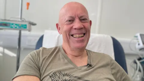 BBC Roy Mitchell, a bald man, smiles broadly from a hospital bed, with medical monitoring equipment around him. He wears a light brown T-shirt.