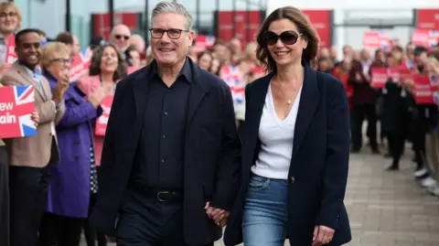 EPA Shutterstock British Prime Minister Sir Keir Starmer and his wife Victoria arrive on the eve of the Labour Party Conference in Liverpool. He wears a dark jacket, shirt and trousers and holds her hand. Victoria wears a navy blue coat, jeans, shades and a white top. 