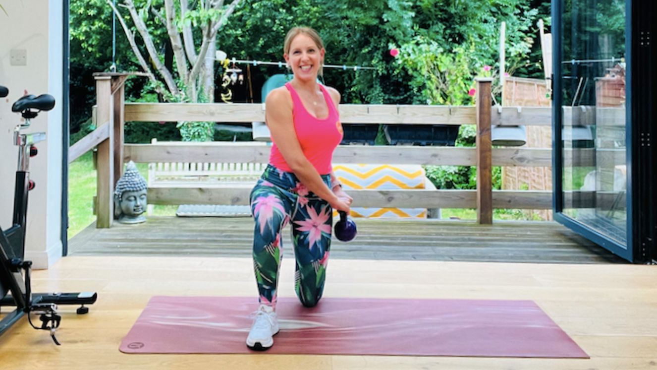 Personal trainer Maddy Biddulph performs a kneeling woodchop. She wears a pink vest, tropical print leggings and white sneakers. She is half-kneeling, with her left knee on the floor and her right leg at a 90 degree angle in front of her. She holds a kettlebell in both hands close to her left hip. Behind her is a leafy garden and next to her is an exercise bike.