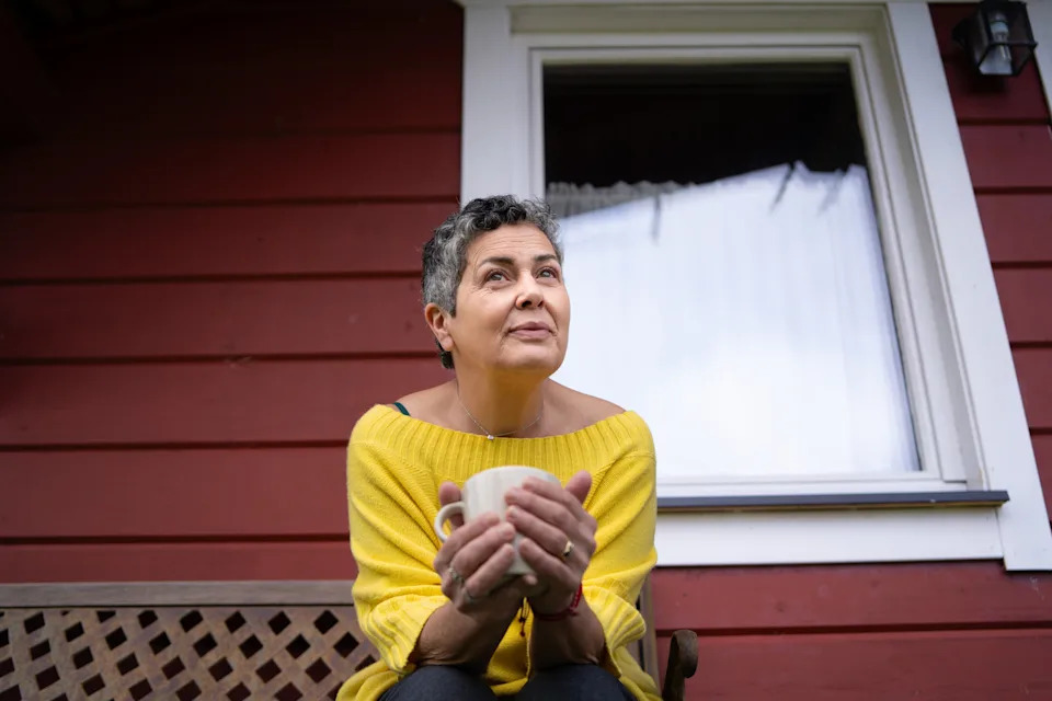 A person sits on a bench outside a red house, wearing an off-shoulder top and holding a mug, looking thoughtful