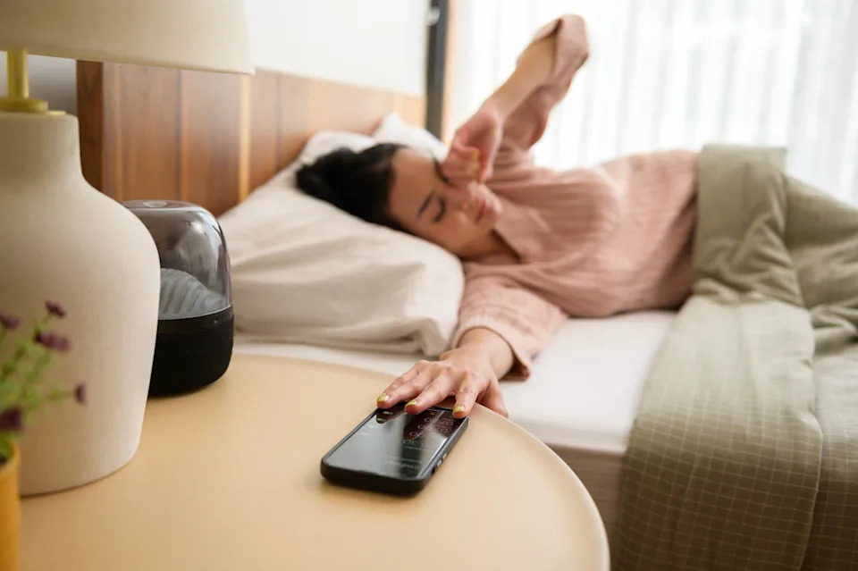Person in bed reaching to turn off smartphone alarm, appearing tired. The scene suggests waking up in the morning