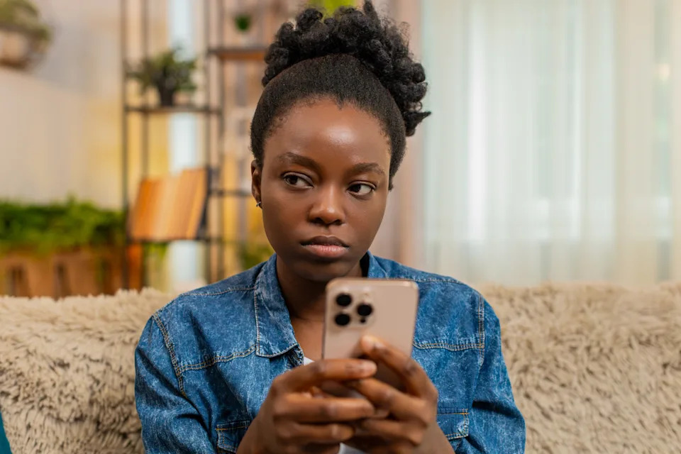 Person sits on a couch, holding a smartphone, appearing thoughtful or concerned, wearing a denim jacket. Living room setting in the background