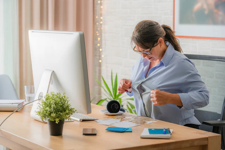 A person at a desk looks at their shirt, searching for stains. The desk has a computer, headphones, a plant, and stationery items