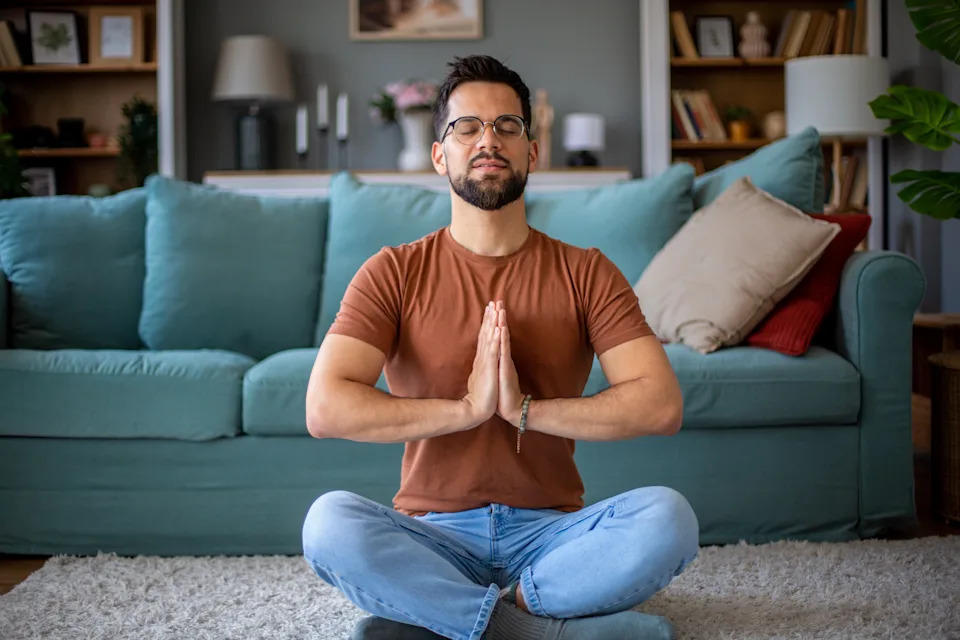 Person meditating in a living room, sitting cross-legged with eyes closed and hands in prayer position. Cozy couch and decor in the background
